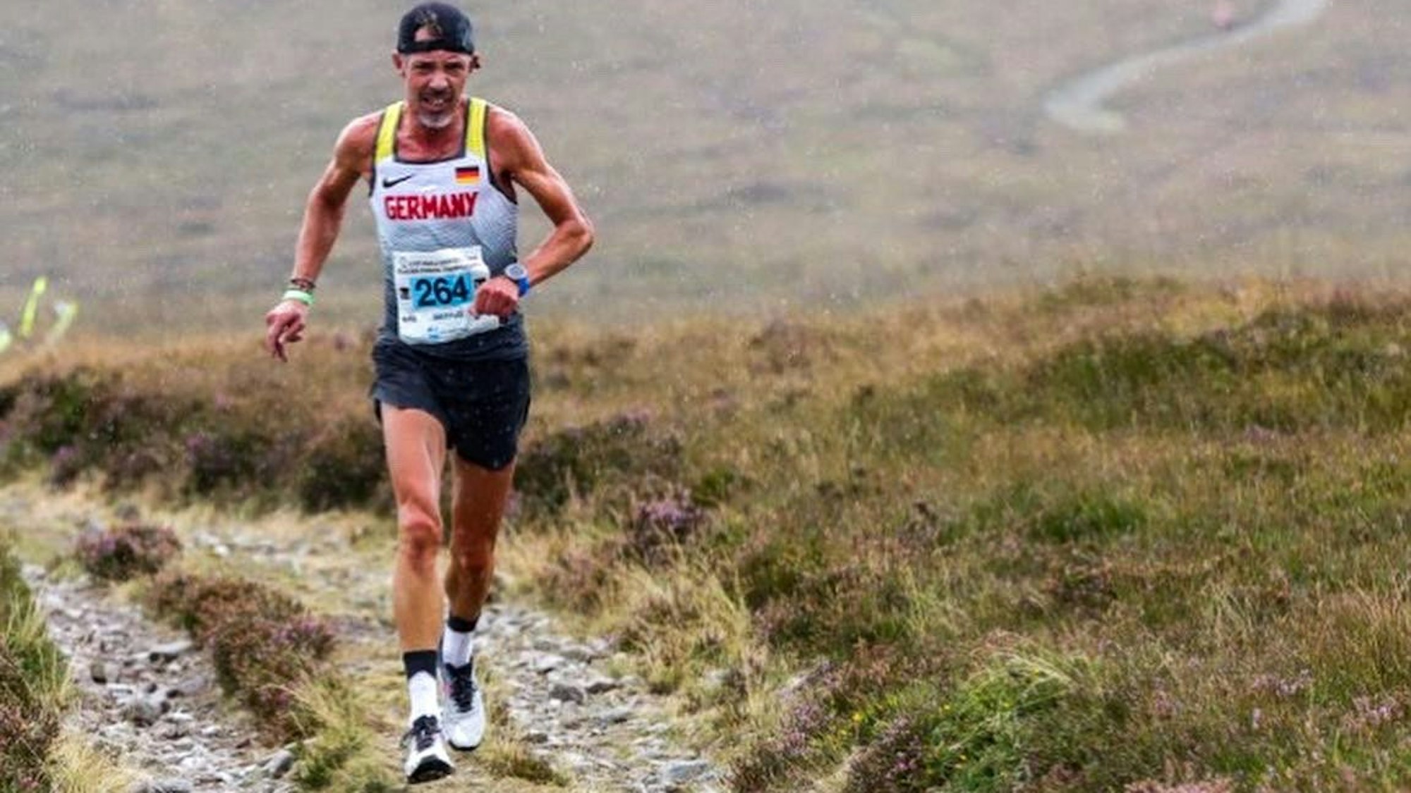 Ein Sportler mit umgekehrt aufgesetzter Kappe läuft über den grasigen Mittelstreifen eines Steinweges im Gebirge.