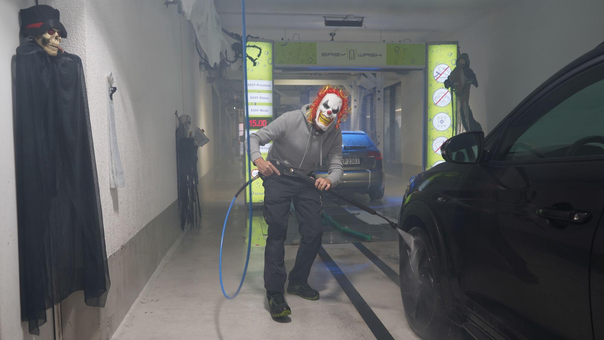 Das Foto zeigt einen Mann mit einer Clown-Maske, der ein Auto wäscht.