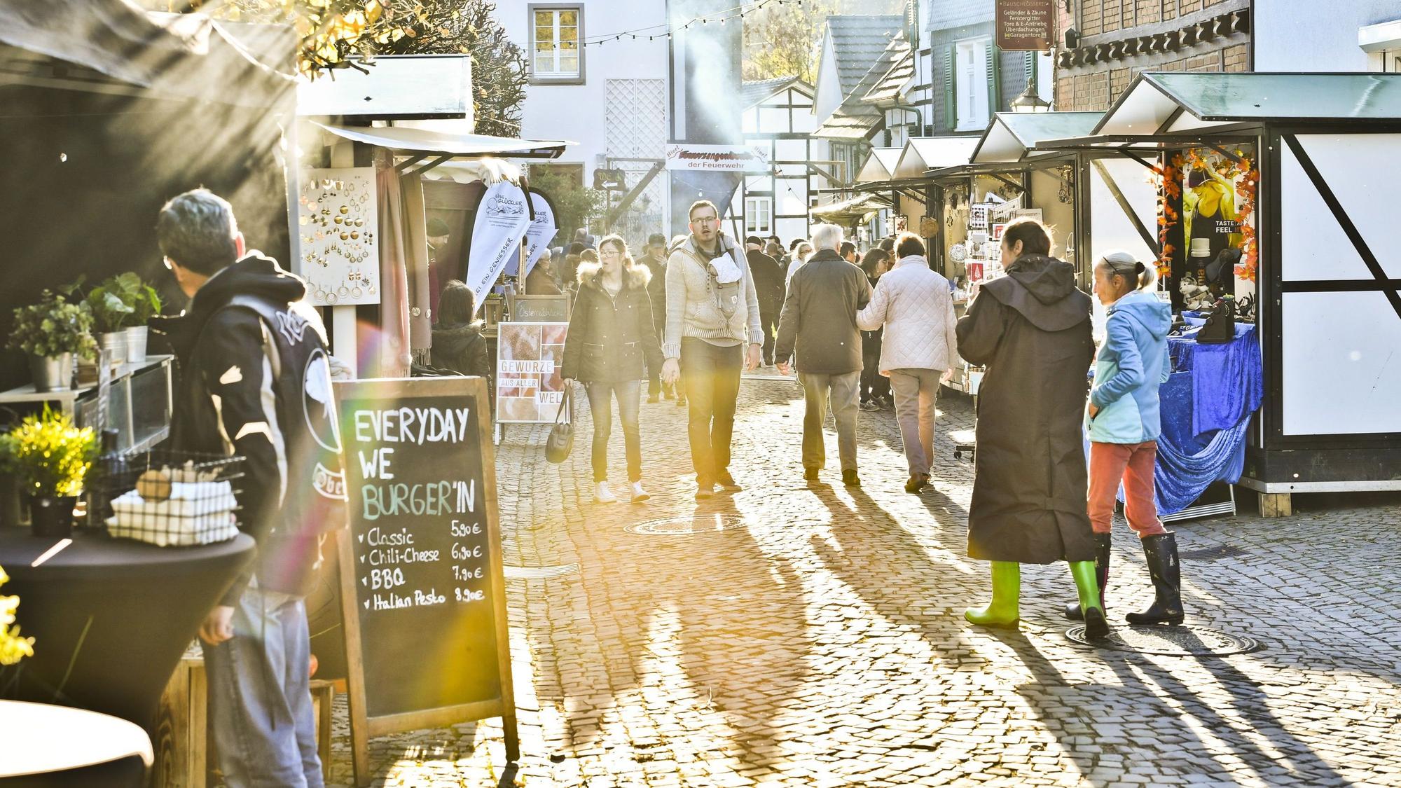 Blick in eine von Buden gesäumte Straße mit Fachwerkhäusern und Kopfsteinpflaster, Menschen schlendern an Verkaufsständen entlang. Sonnenstrahlen tauchen die Szene in warmes Licht.