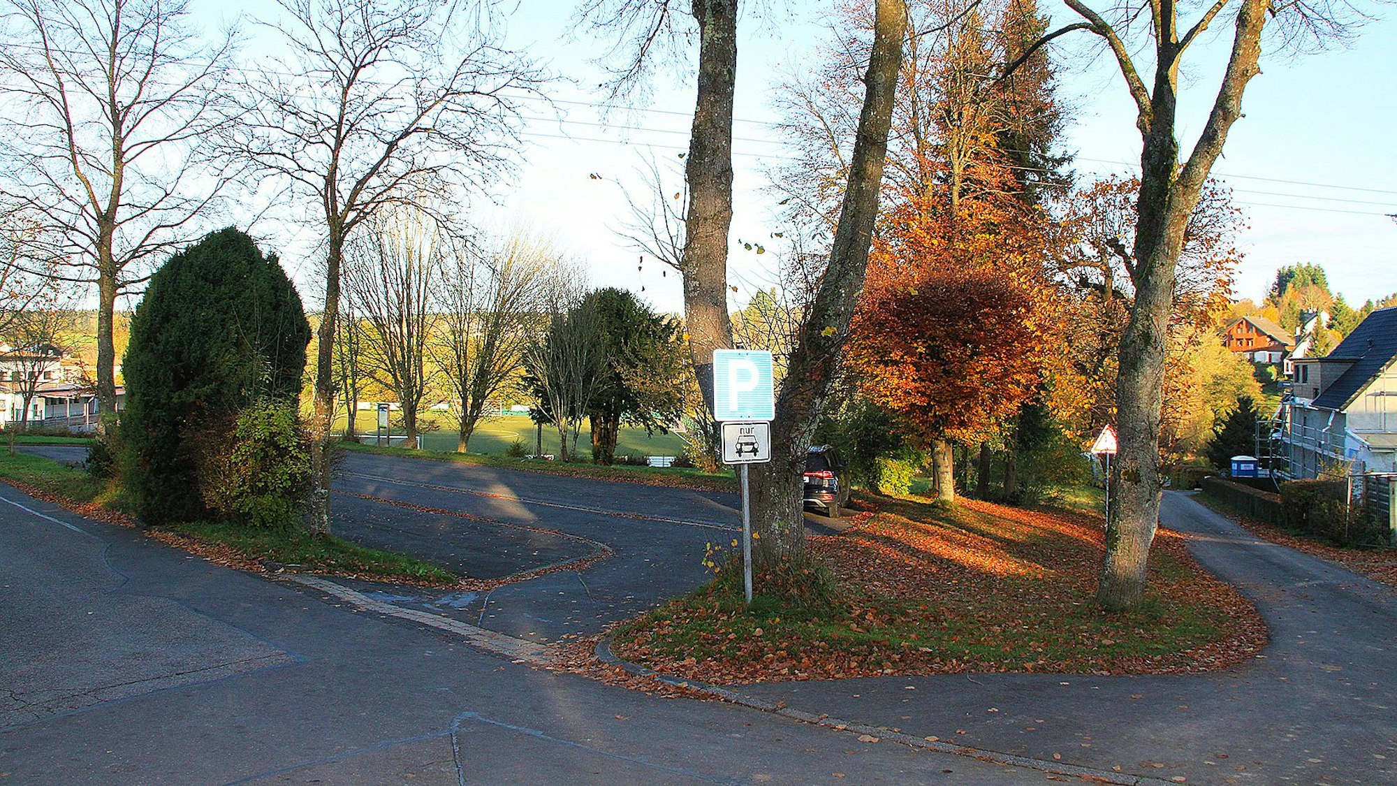 Nahe des Sportplatzes in Dahlem-Schmidtheim ist ein kleiner Parkplatz, der in einem schlechten Zustand ist. Rund um den asphaltierten Platz sind Bäume zu sehen.