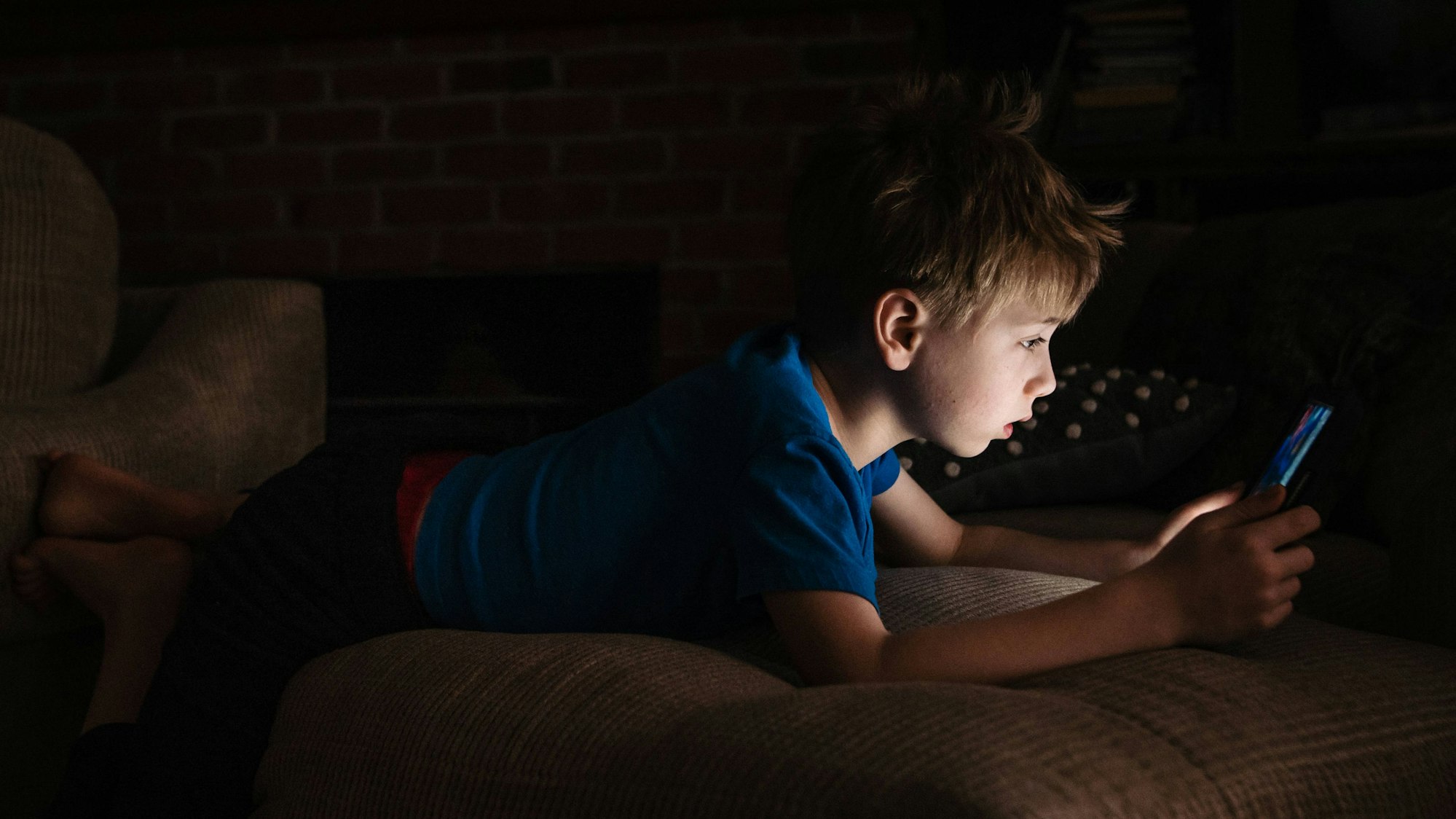 Ein Junge liegt in einem dunklen Raum auf einem Sofa und schaut ängstlich auf seinen Tablet-Bildschirm.