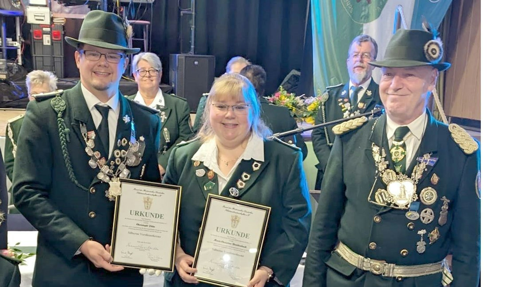Eine Frau und zwei Männer in dunkelgrünen Uniformen lächeln, zwei halten Urkunden in Bilderrahmen in den Händen.