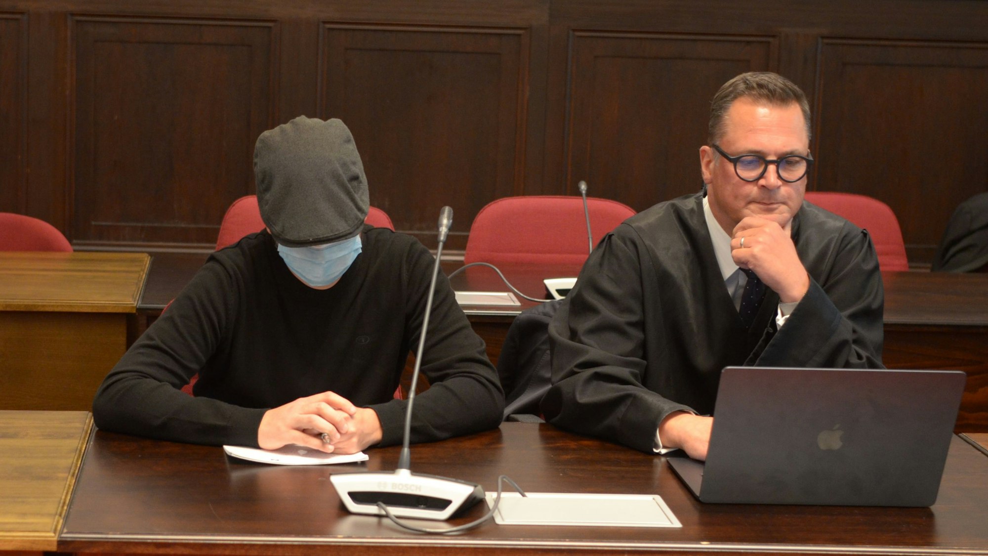 Einer der Angeklagten sitzt mit geneigtem Kopf, Schutzmaske und Kappe neben seinem Verteidiger.