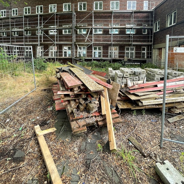 Die Hinterseite eines dunklen Backsteinbaus ist zu sehen, mit Gerüst und einigen Baubrettern und Bauzaun.