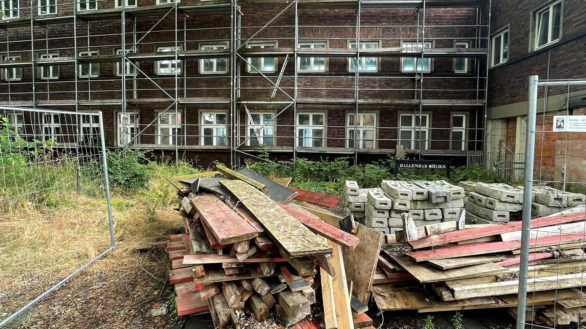 Die Hinterseite eines dunklen Backsteinbaus ist zu sehen, mit Gerüst und einigen Baubrettern und Bauzaun.