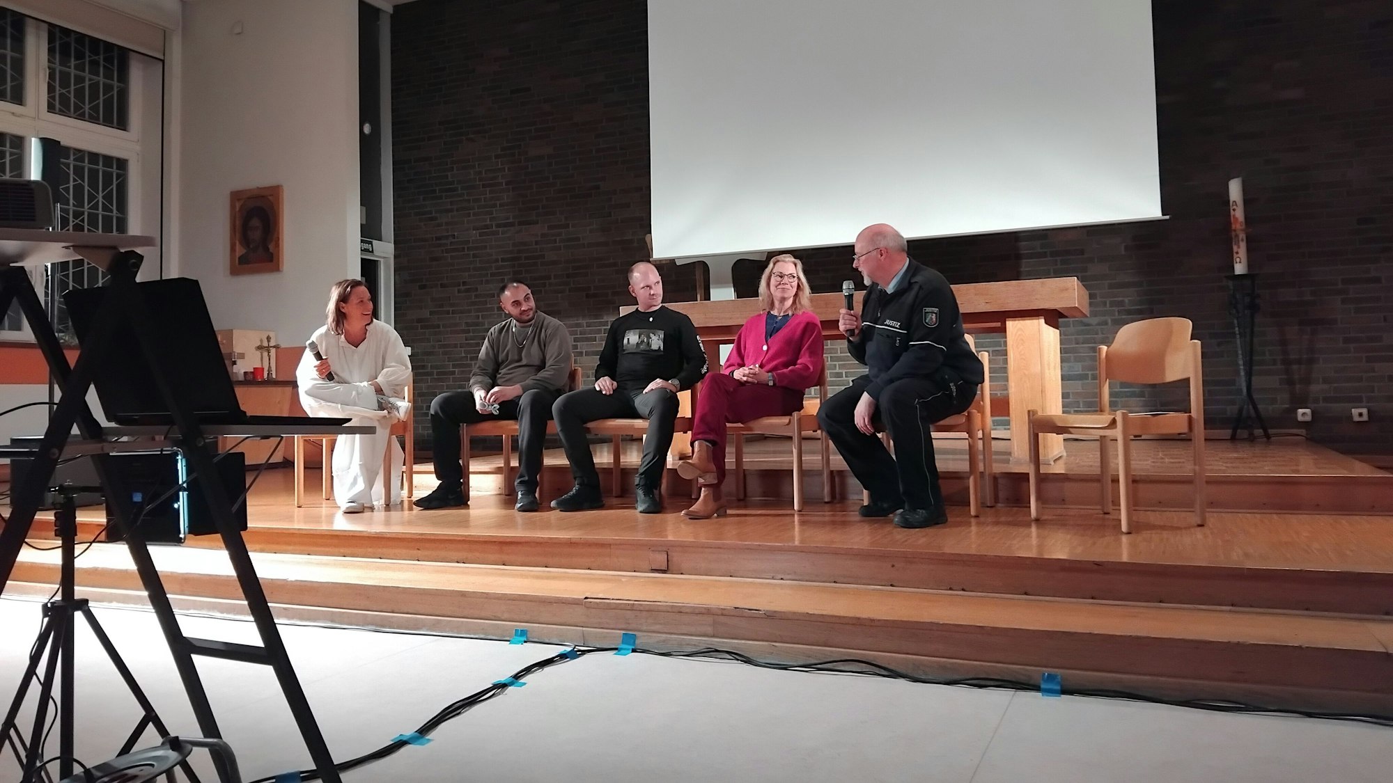 Fünf Leute sitzen auf Stühlen vor einer Leinwand, ein Mann hält ein Mikrophon, im Vordergrund sieht man eine Kamera.