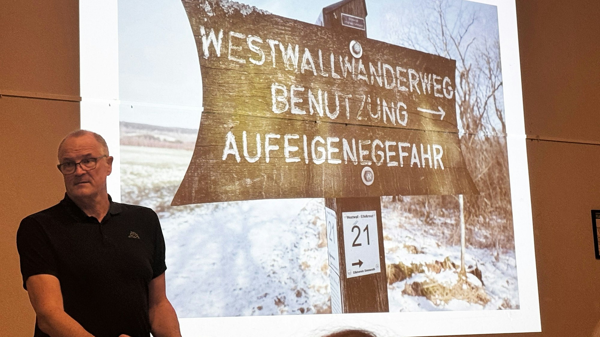 Ein Mann redet zum Publikum, hinter ihm ist ein Foto mit der Aufschrift „Westwallwanderweg - Benutzung auf eigene Gefahr“ zu sehen.