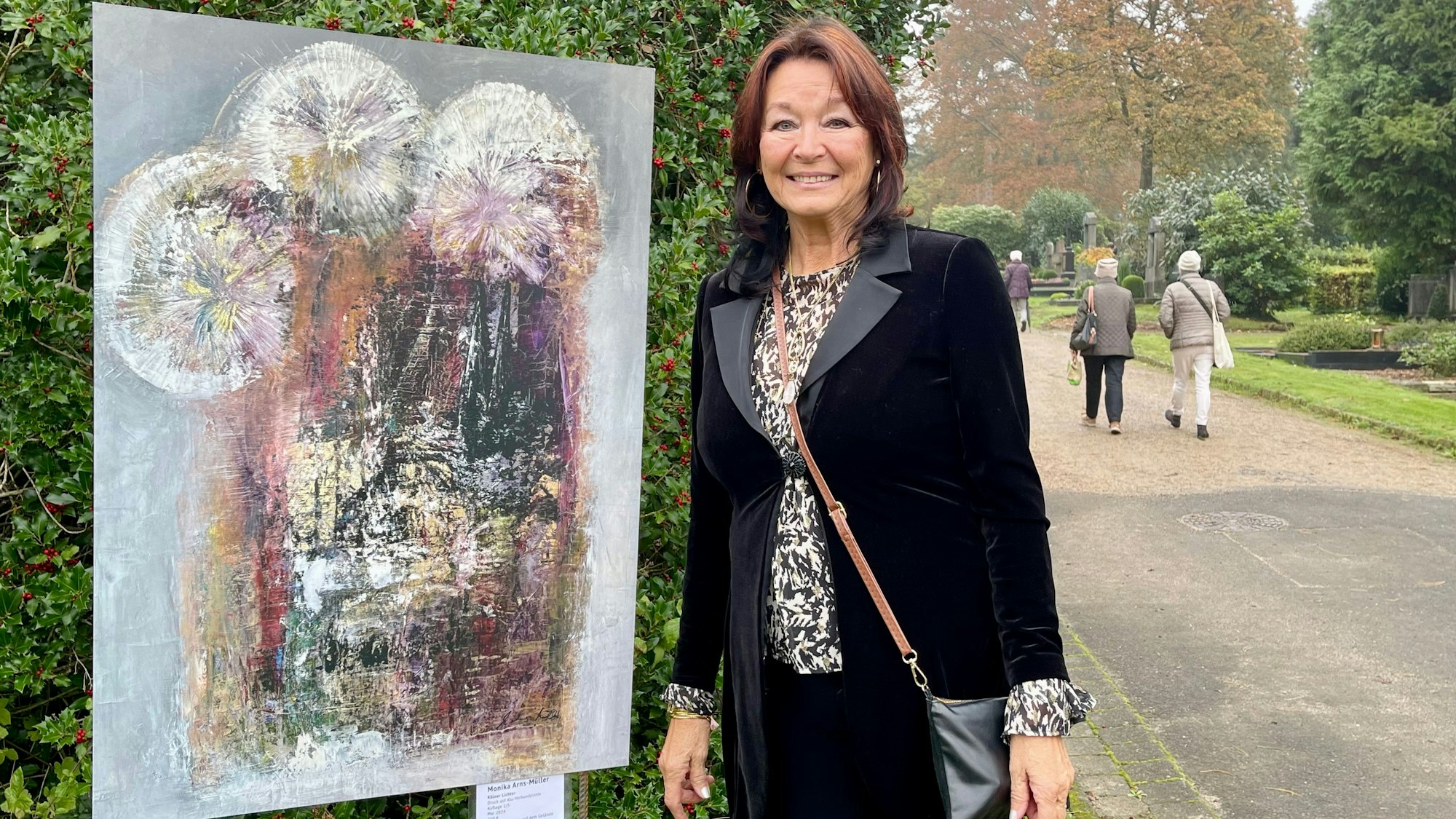 Eine Frau steht neben einem Bild, das eine stilisierte und farbige Darstellung des Kölner Doms mit Feuerwerk zeigt.Im Hintergrund sind Gräber und Friedhofsbesucher.