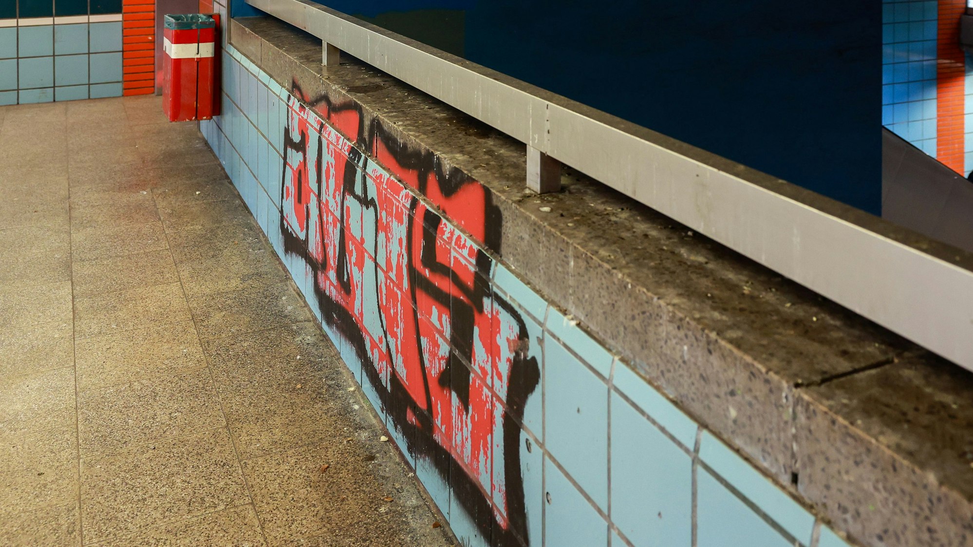 Graffiti auf den bunten Kacheln an einer Brüstung im Zwischengeschoss der U-Bahn-Haltestelle Geldernstraße/Parkgürtel. Auf dem Boden Brotkrumen als Taubenfutter
