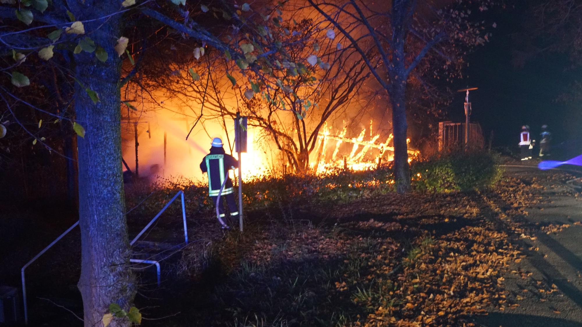 Ein Feuerwehrmann steht nachts vor einer brennenden Gartenhütte und löscht diese mit Wasser aus einem Strahlrohr.