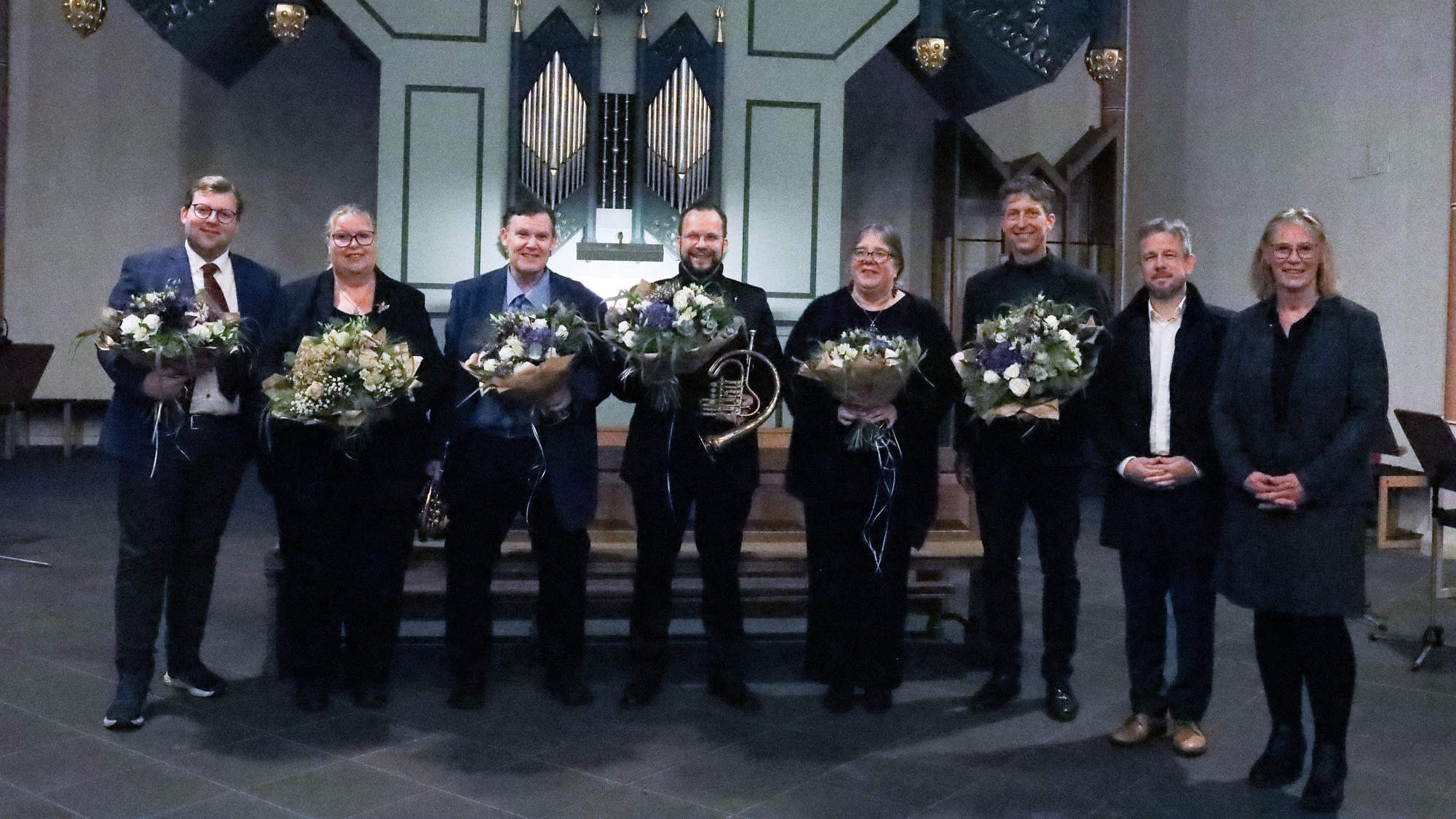 Acht Menschen schauen in Richtung der Kamera, sechs von ihnen halten Blumensträuße.