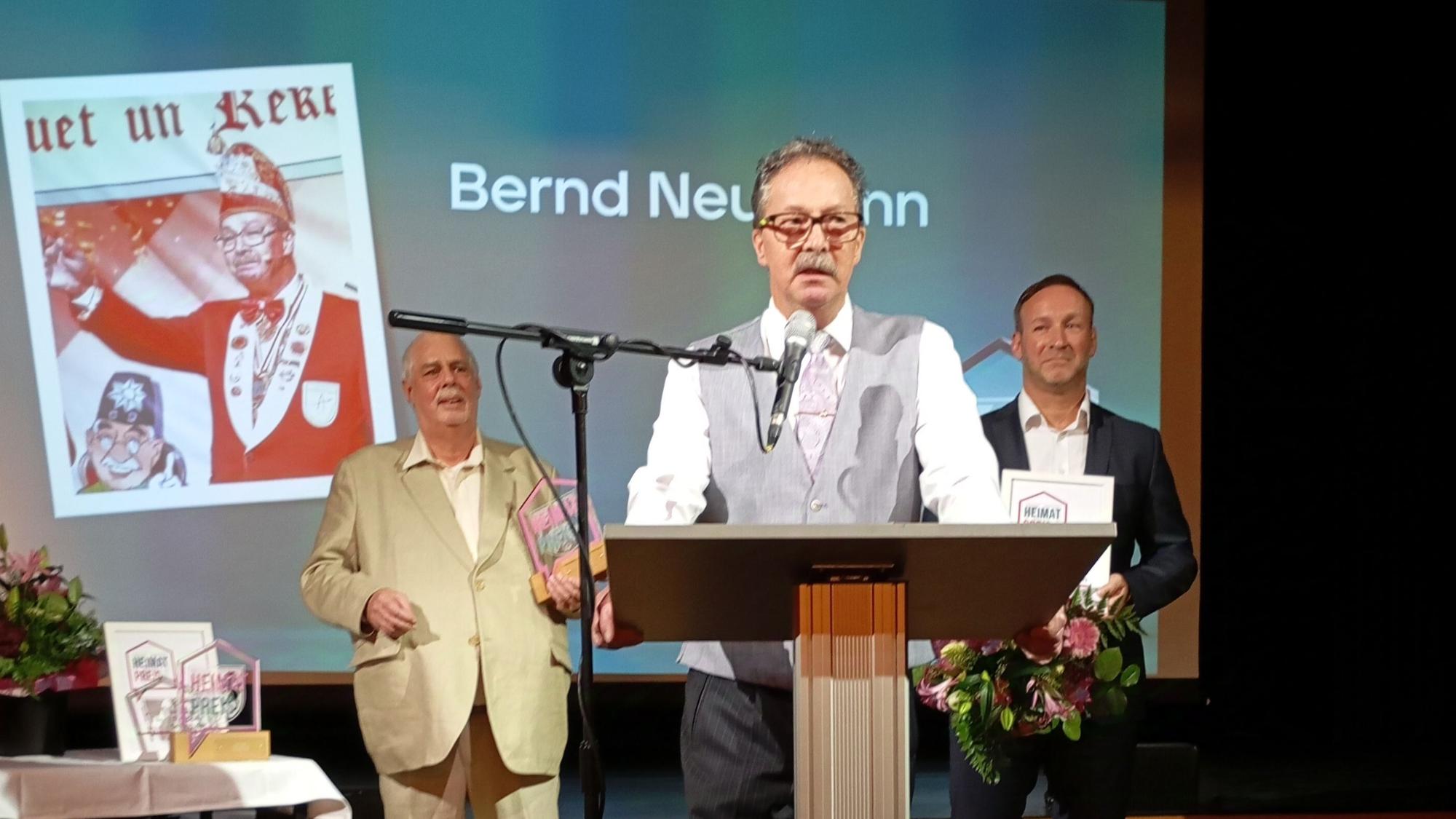 Bernd Neumann trägt eine graue Weste und ein weißes Hemd. Er steht an einem Pult und spricht in ein Mikrofon.