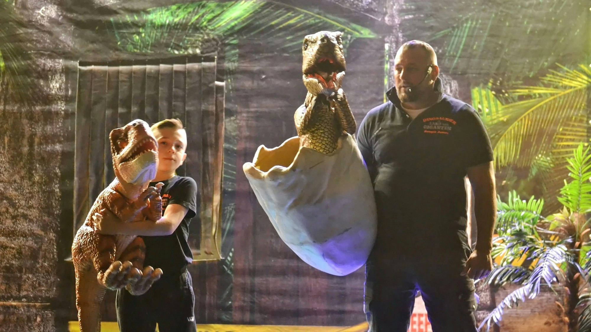 Ein Mann und ein Junge präsentieren Dinosaurier-Nachbildungen auf einer Bühne mit Urwald-Ästhetik.
