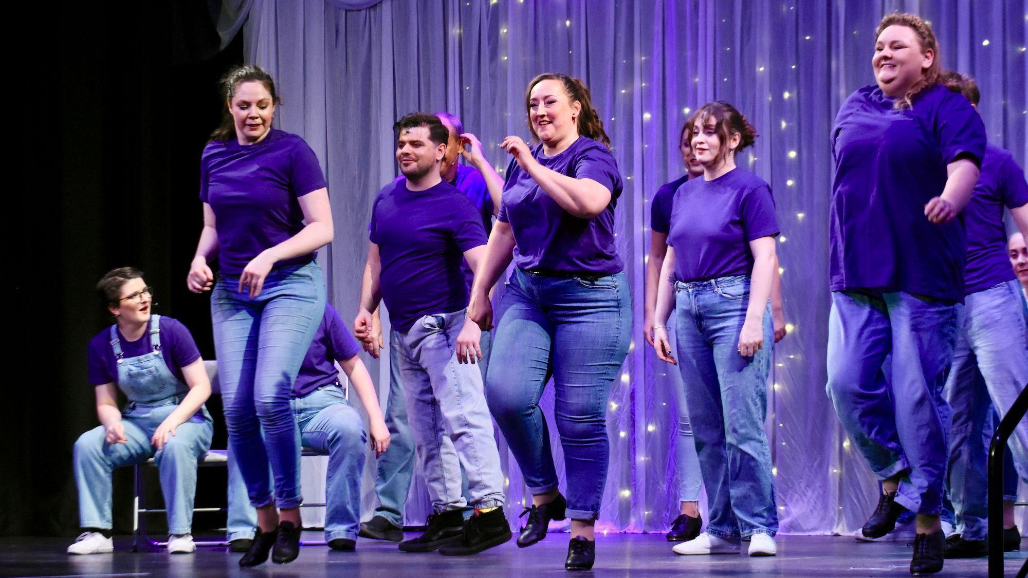 Frauen und Männer in Jeans und blauen Oberteilen tanzen auf einer Bühne.