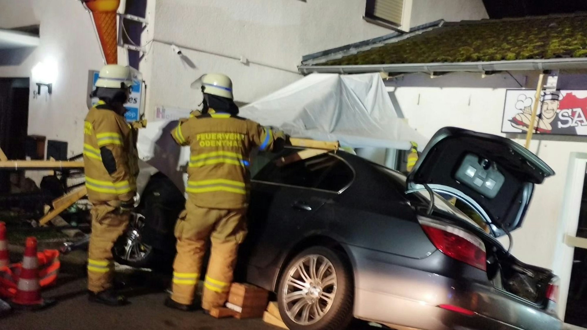 Zwei Feuerwehrleute stehen neben einem Unfallauto vor einem Gebäude mit einem Eiscafé-Schild.