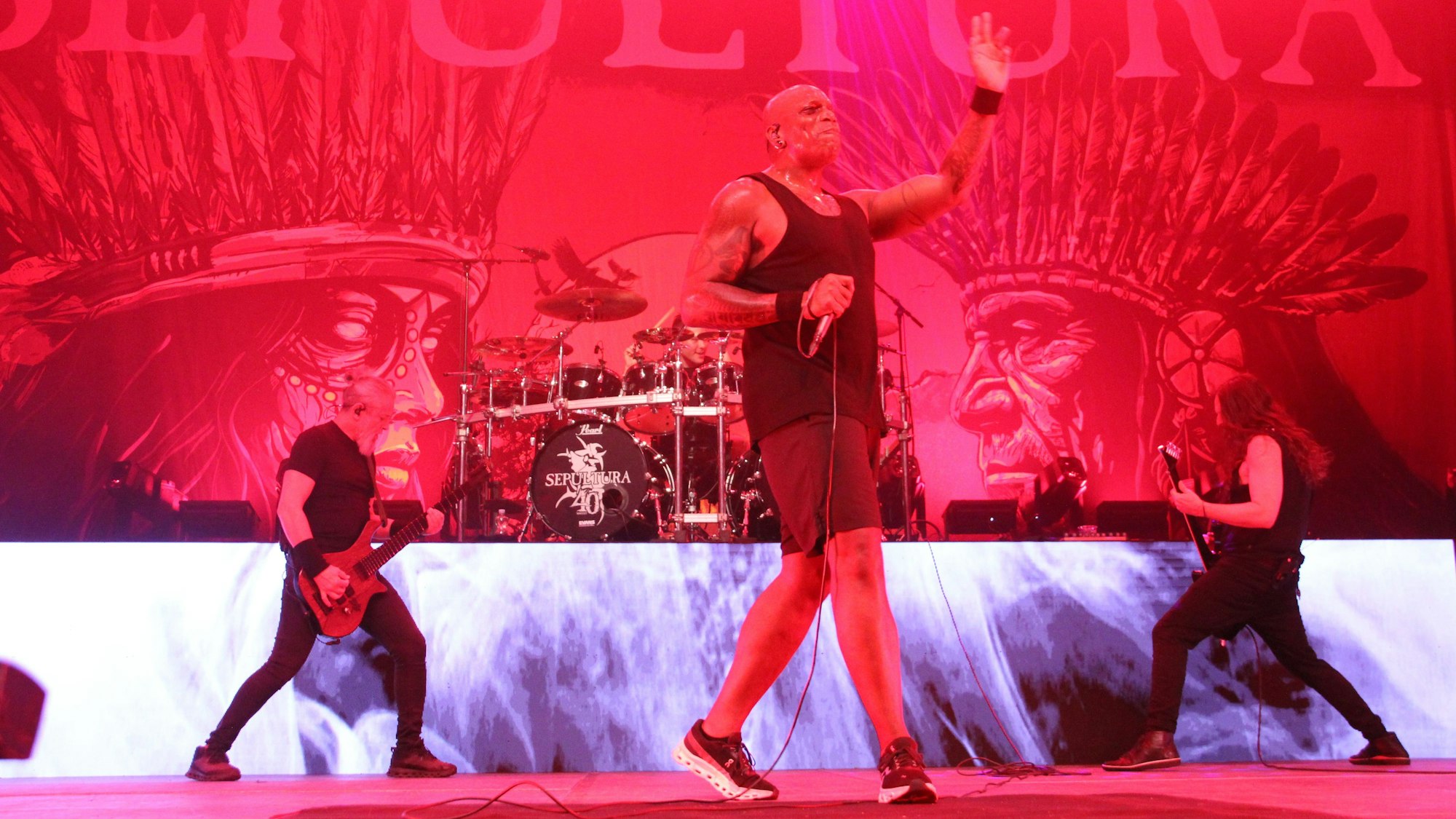 Zwei Gitarristen umrahmen den Sänger der Band. Im Hintergrund thront das Schlagzeug auf einem Podest.