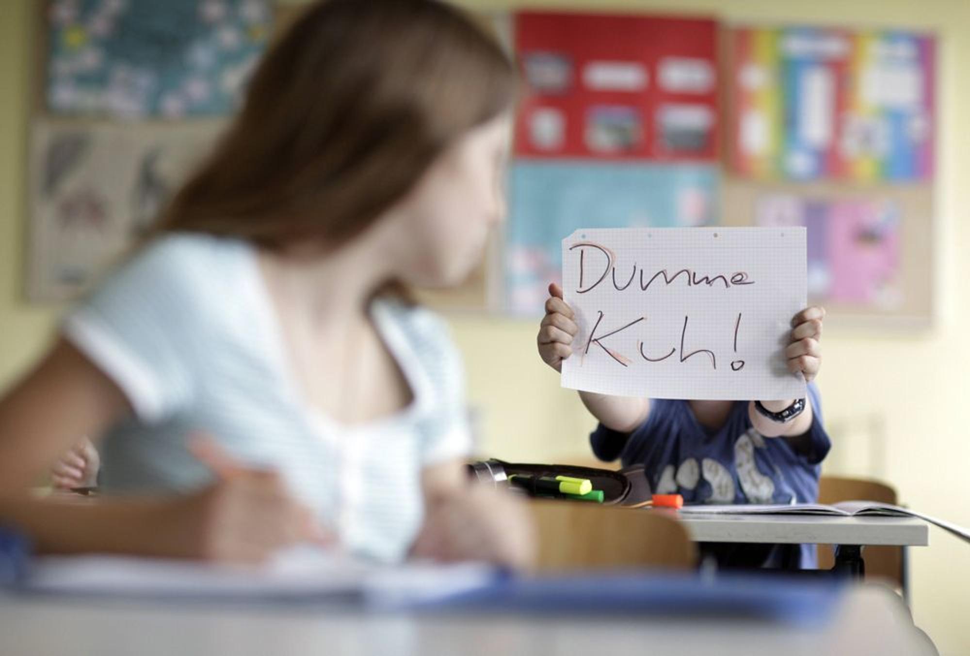 Das Bild zeigt ein junges Mädchen, das von einem Mitschüler gemobbt wird. Er hält ihr ein Schild mit der Aufschrift „Dumme Kuh!“ entgegen. (Symbolbild)