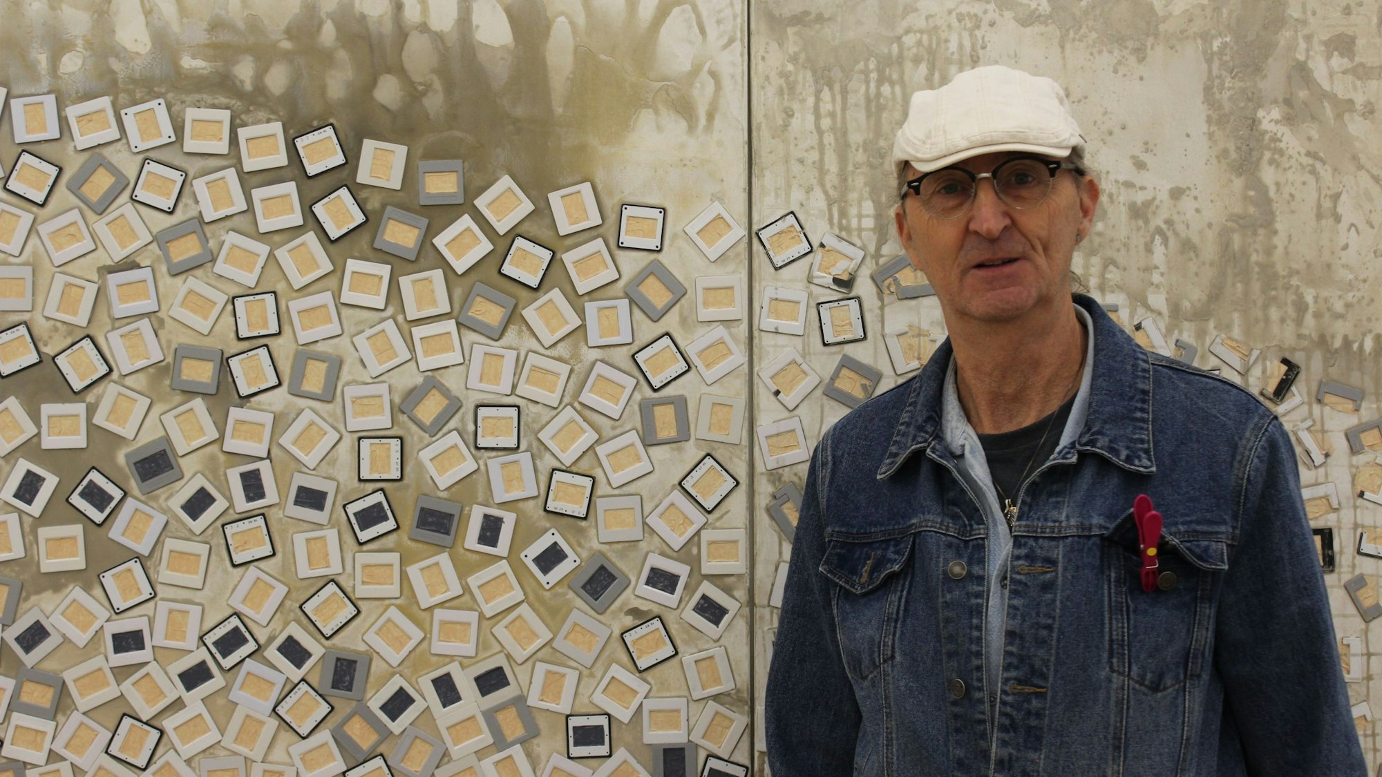 Bernhard Johannes Trier steht vor seinem großen Kunstwerk an der Wand. Er trägt eine Jeansjacke und eine weiße Mütze.