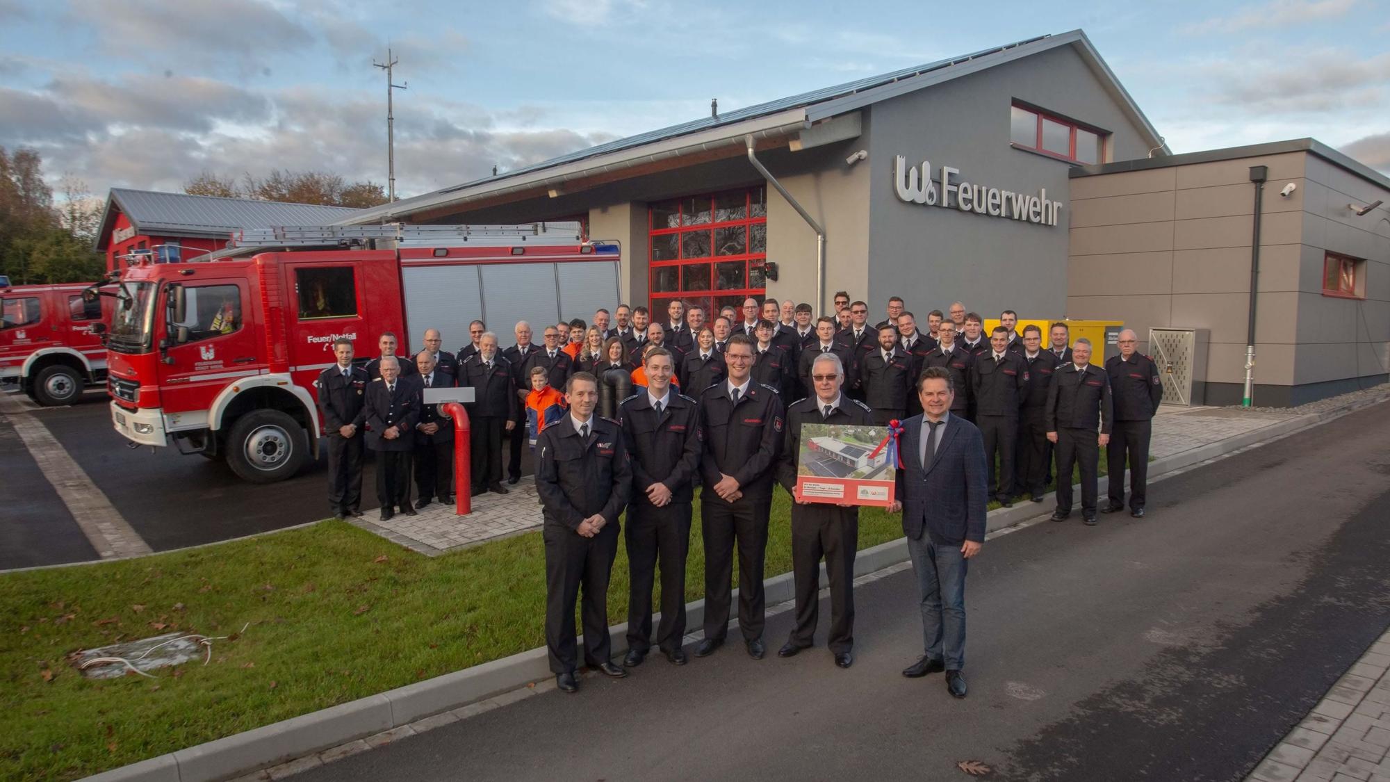 Eine Gruppe Feuerwehrleute und ein Bürgermeister stehen vor einem Feuerwehrhaus mit neuem Anbau.