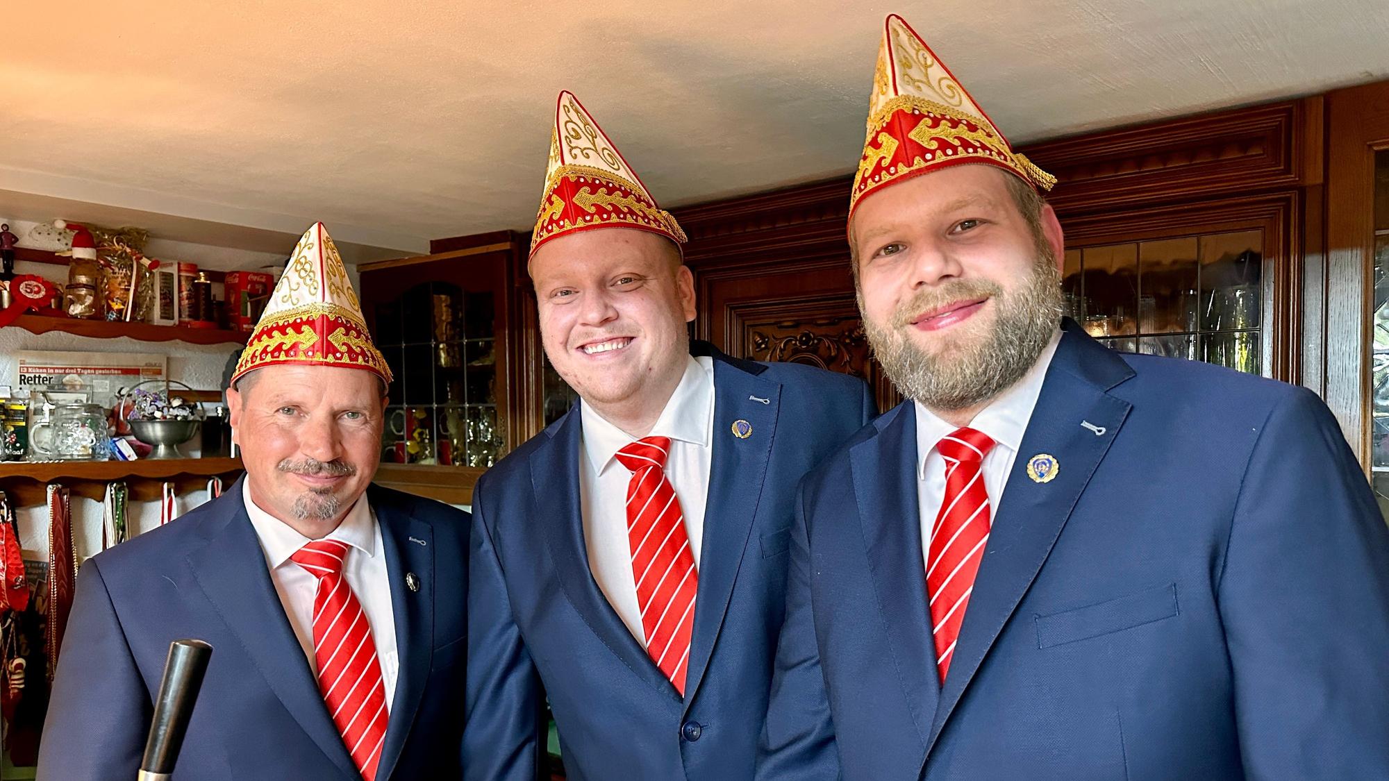 Drei Männer in identischen blauen Anzügen mit rot-weiß-gestreiften Krawatten und Schiffchen lachen in die Kamera.