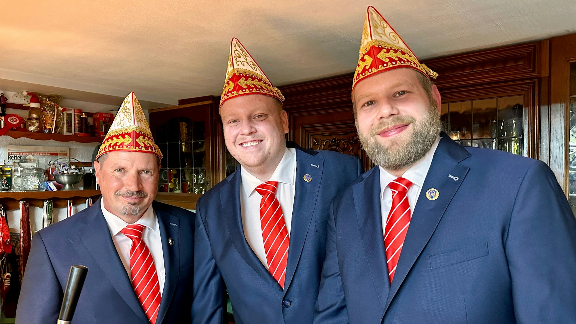 Drei Männer in identischen blauen Anzügen mit rot-weiß-gestreiften Krawatten und Schiffchen lachen in die Kamera.