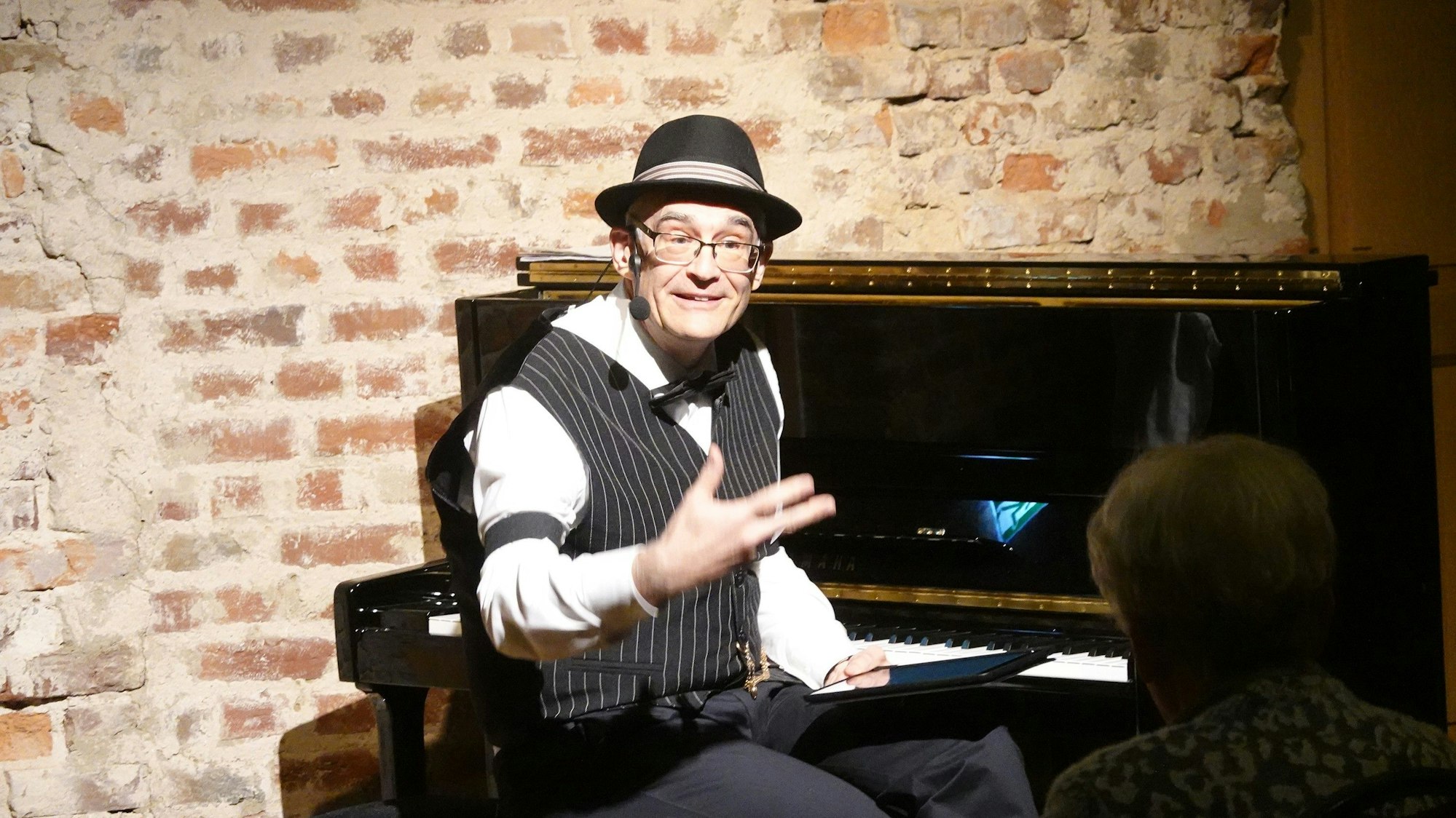 Der Musiker träg ein weißes Hemd, eine schwarze Weste und einen Hut. Er sitzt an einem Klavier.