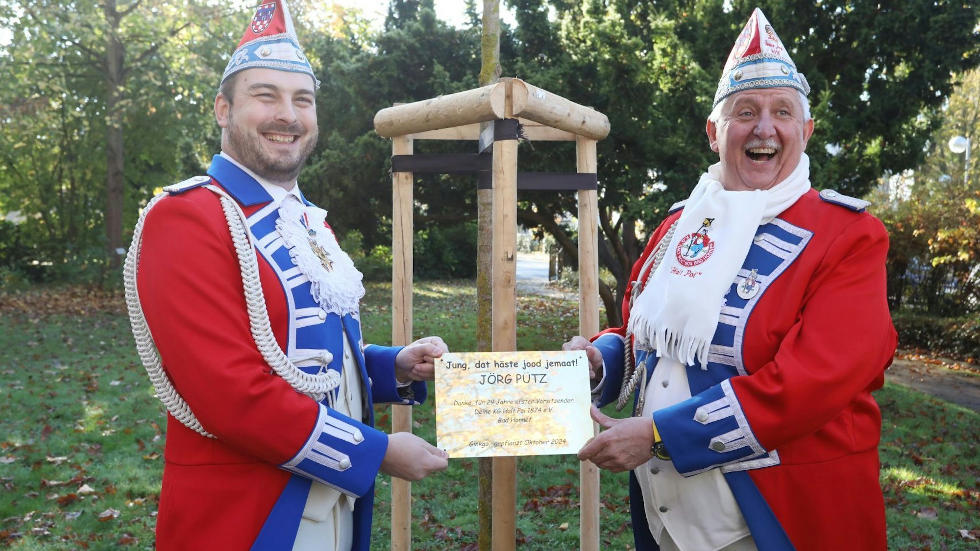 Zwei Männer in rot-weiß-blauer Gesellschaftskleidung der KG Halt Pol halten ein kleines goldenes Schild in den Händen.
