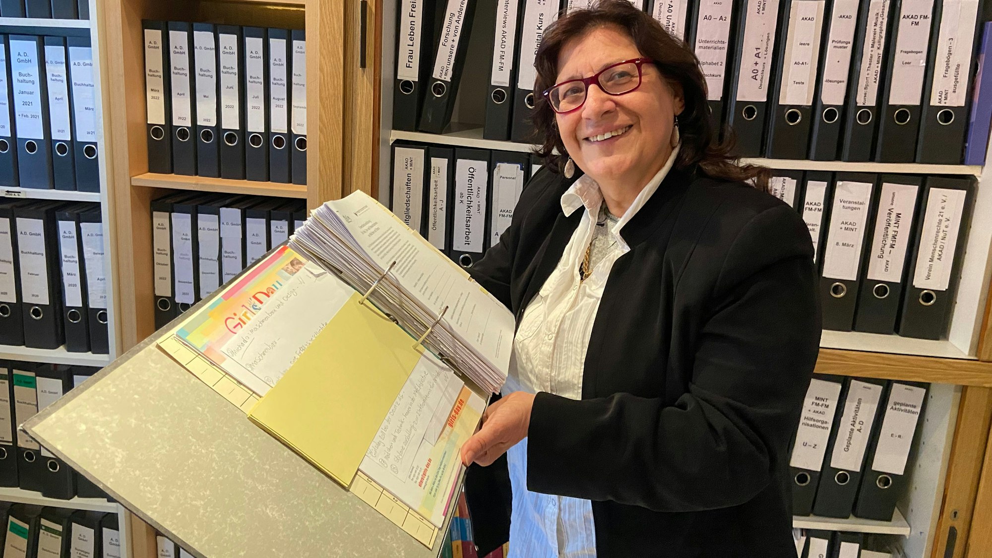 Eine Frau mit dunklen Haaren und Brille hält einen aufgeklappten Aktenordner. Sie steht vor einem Regal mit Aktenordnern.