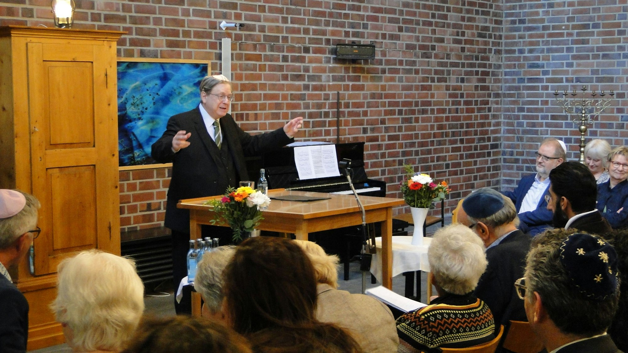 Ein Mann im dunklen Anzug singt mit ausgebreiteten Armen vor vielen sitzenden Menschen, die zum Teil Kippa tragen.