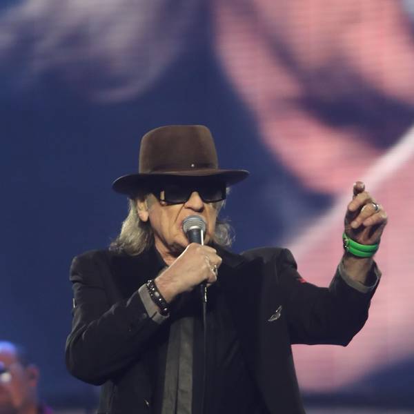 Udo Lindenberg, mit Hut und dunkler Brille, singt mit Mikro in der rechten Hand während eines Konzerts im Juni 2022 in der Kölner Lanxess Arena. Die linke Hand hat er erhoben; ein grünes Armband schmückt sein Handgelenk.