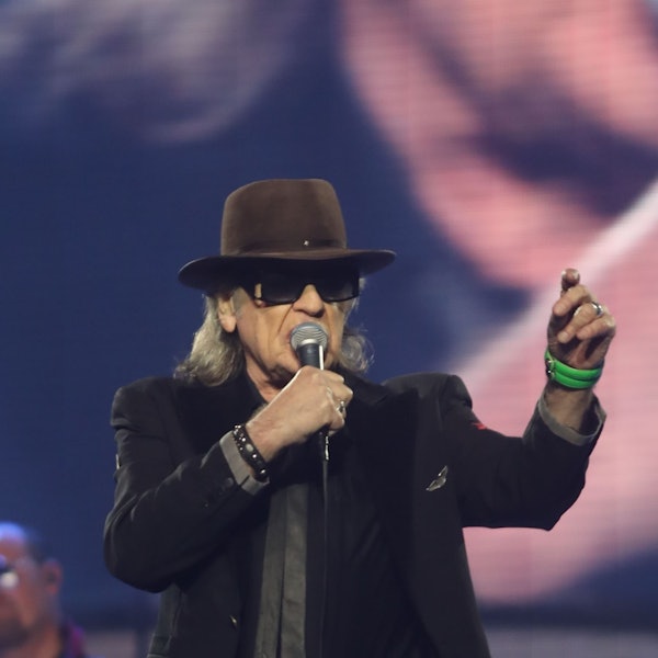 Udo Lindenberg, mit Hut und dunkler Brille, singt mit Mikro in der rechten Hand während eines Konzerts im Juni 2022 in der Kölner Lanxess Arena. Die linke Hand hat er erhoben; ein grünes Armband schmückt sein Handgelenk.