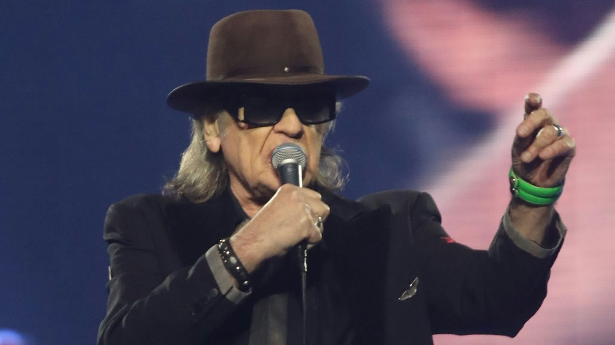 Udo Lindenberg, mit Hut und dunkler Brille, singt mit Mikro in der rechten Hand während eines Konzerts im Juni 2022 in der Kölner Lanxess Arena. Die linke Hand hat er erhoben; ein grünes Armband schmückt sein Handgelenk.