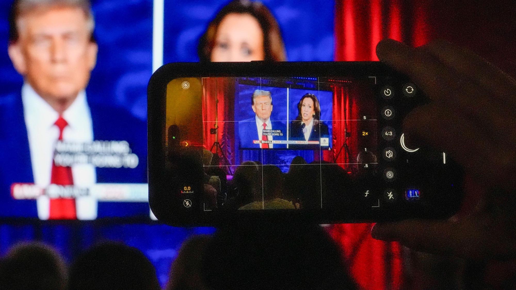 Eine Person filmt mit dem Handy den Bildschirm, auf dem eine Debatte zwischen Donald Trump und Kamala Harris übertragen wird.