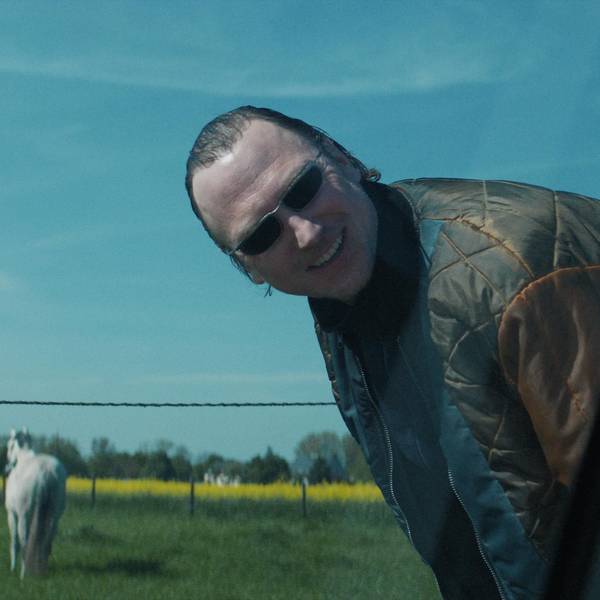 Der Ganove Johnny trägt eine Sonnenbrille und schaut – an einer Straße stehend – lachend in Richtung einer Autofensterscheibe.