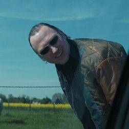 Der Ganove Johnny trägt eine Sonnenbrille und schaut – an einer Straße stehend – lachend in Richtung einer Autofensterscheibe.