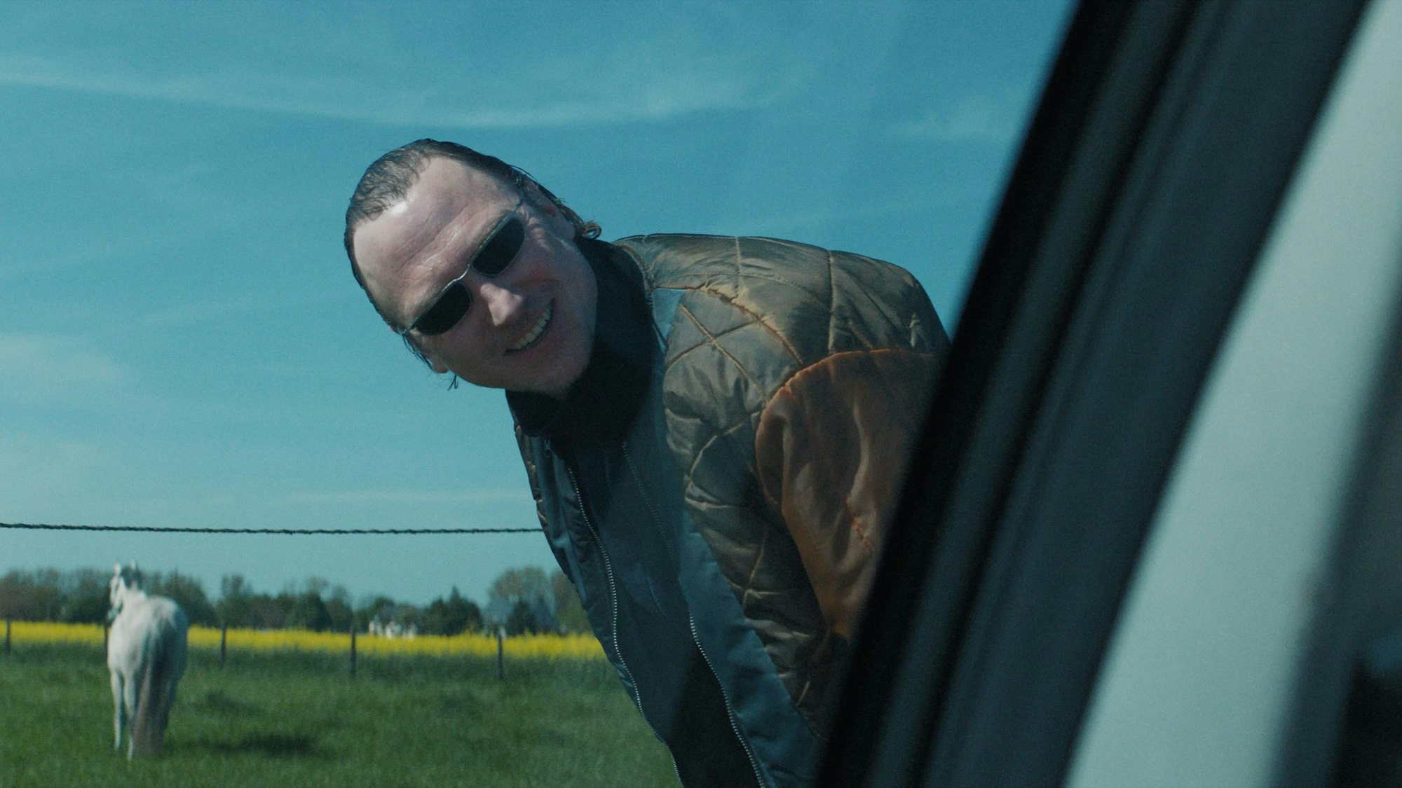 Der Ganove Johnny trägt eine Sonnenbrille und schaut – an einer Straße stehend – lachend in Richtung einer Autofensterscheibe.