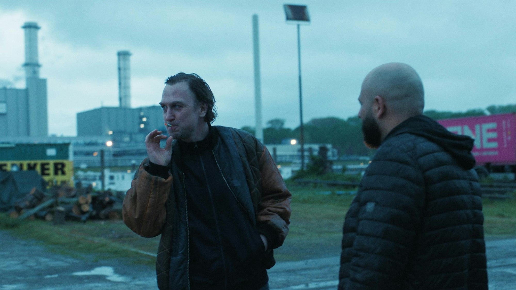 Johnny (links im Bild) raucht eine Zigarette, schaut dabei nach links. Rechts neben ihm steht ein Mann, den Rücken zur Kamera gewandt. Im Hintergrund Leverkusener Chemiebauten.