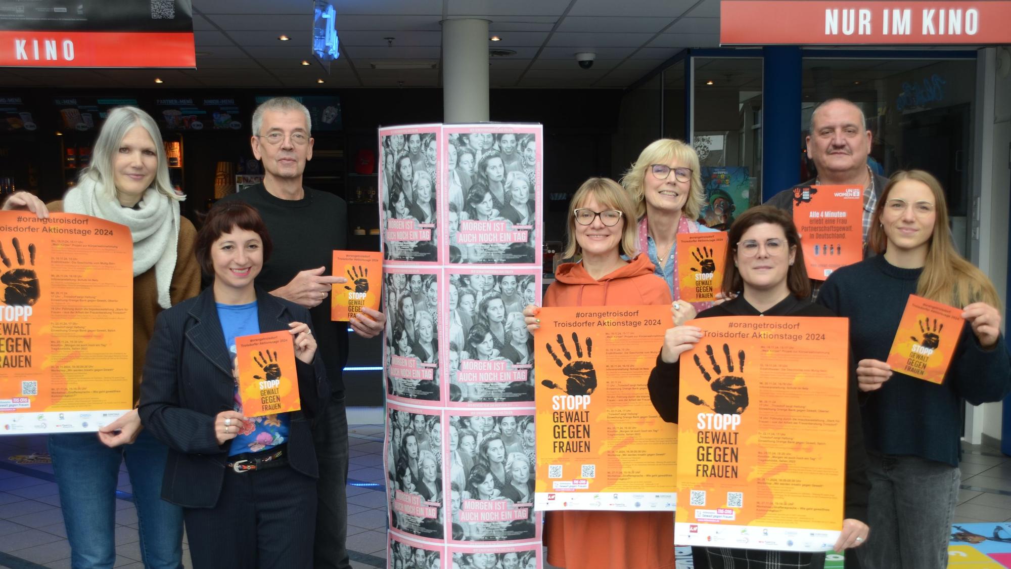 Eine Gruppe von sechs Frauen und zwei Männern hat sich zum Gruppenfoto aufgestellt. Sie tragen orange Plakate mit einer schwarzen zum Stopp gespreizten Handfläche.
