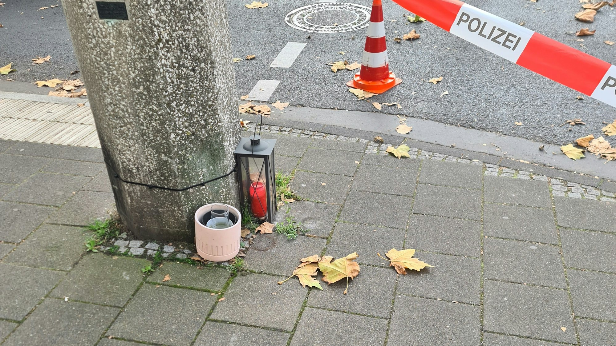 Zu sehen ist Grabschmuck, der an einer Säule auf einem Bürgersteig steht. Zu sehen ist auch rot-weißes Absperrband mit der Aufschrift „Polizei“.