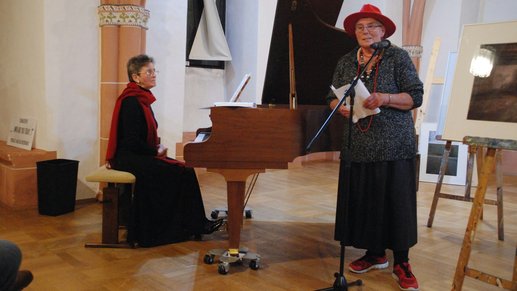 Eine Frau sitzt an einem Flügel, während eine andere Frau Zettel in den Händen hält und hinter einem Mikrofonständer steht.