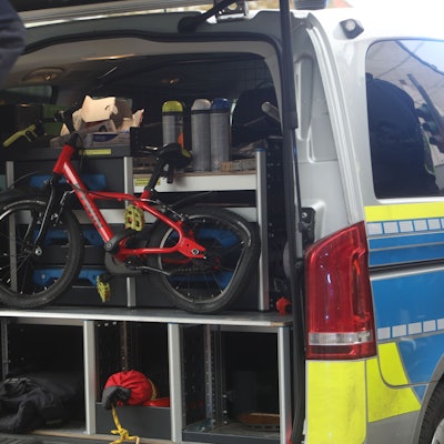 Zu sehen ist ein rotes Kinderfahrrad, dessen Hinterrad kaputt ist. Es befindet sich im Kofferraum eines Einsatzfahrzeugs der Kölner Polizei. Auf der linken Seite ist ein Polizist zu sehen.