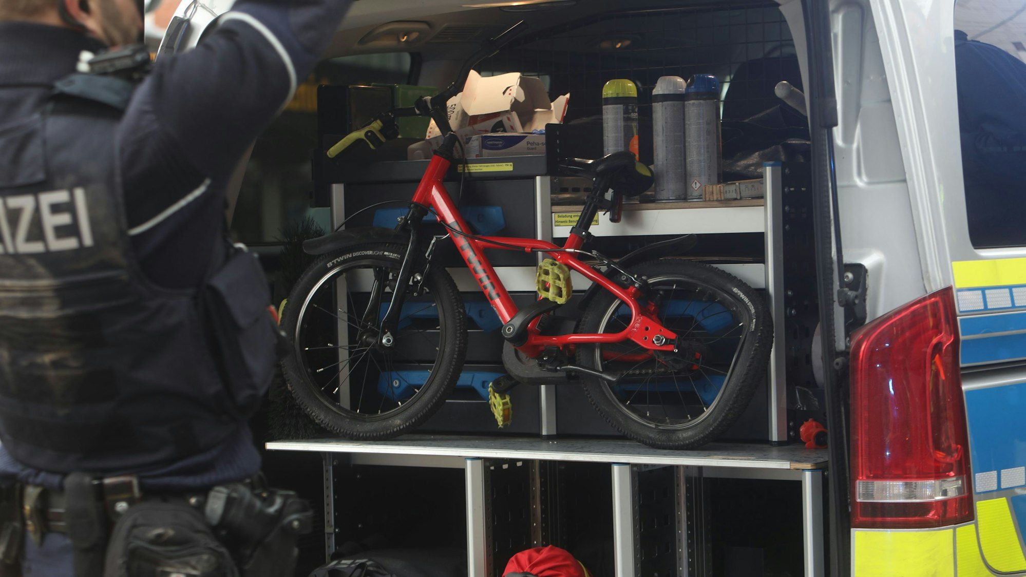 Zu sehen ist ein rotes Kinderfahrrad, dessen Hinterrad kaputt ist. Es befindet sich im Kofferraum eines Einsatzfahrzeugs der Kölner Polizei. Auf der linken Seite ist ein Polizist zu sehen.