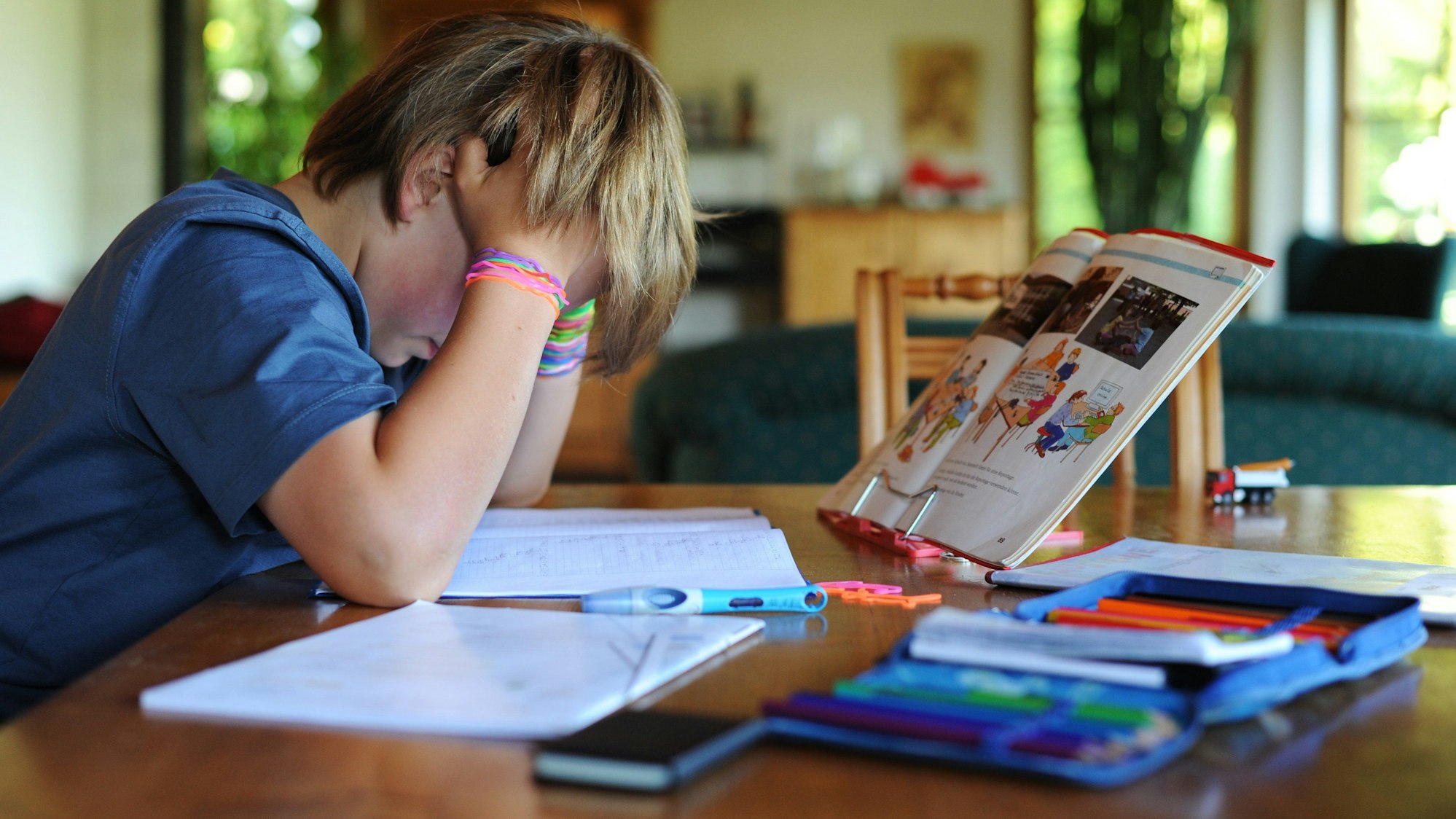 Ein Junge hält sich beim Erledigen seiner Hausaufgaben den Kopf. Vor und neben ihm liegen Schulhefte.