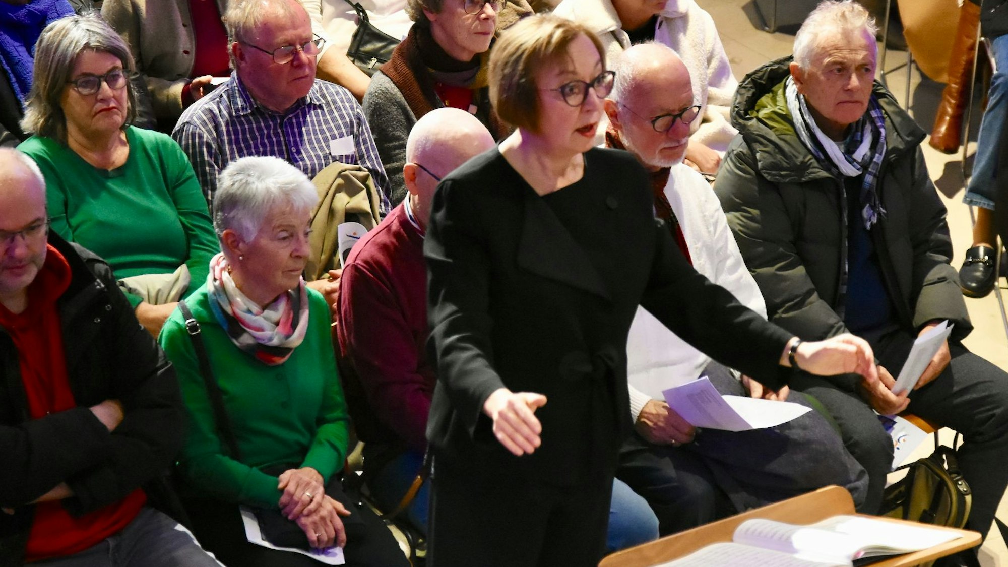 Eine Frau steht an einem Dirigentenpult, hinter ihr sitzen Männer und Frauen, die zuhören.