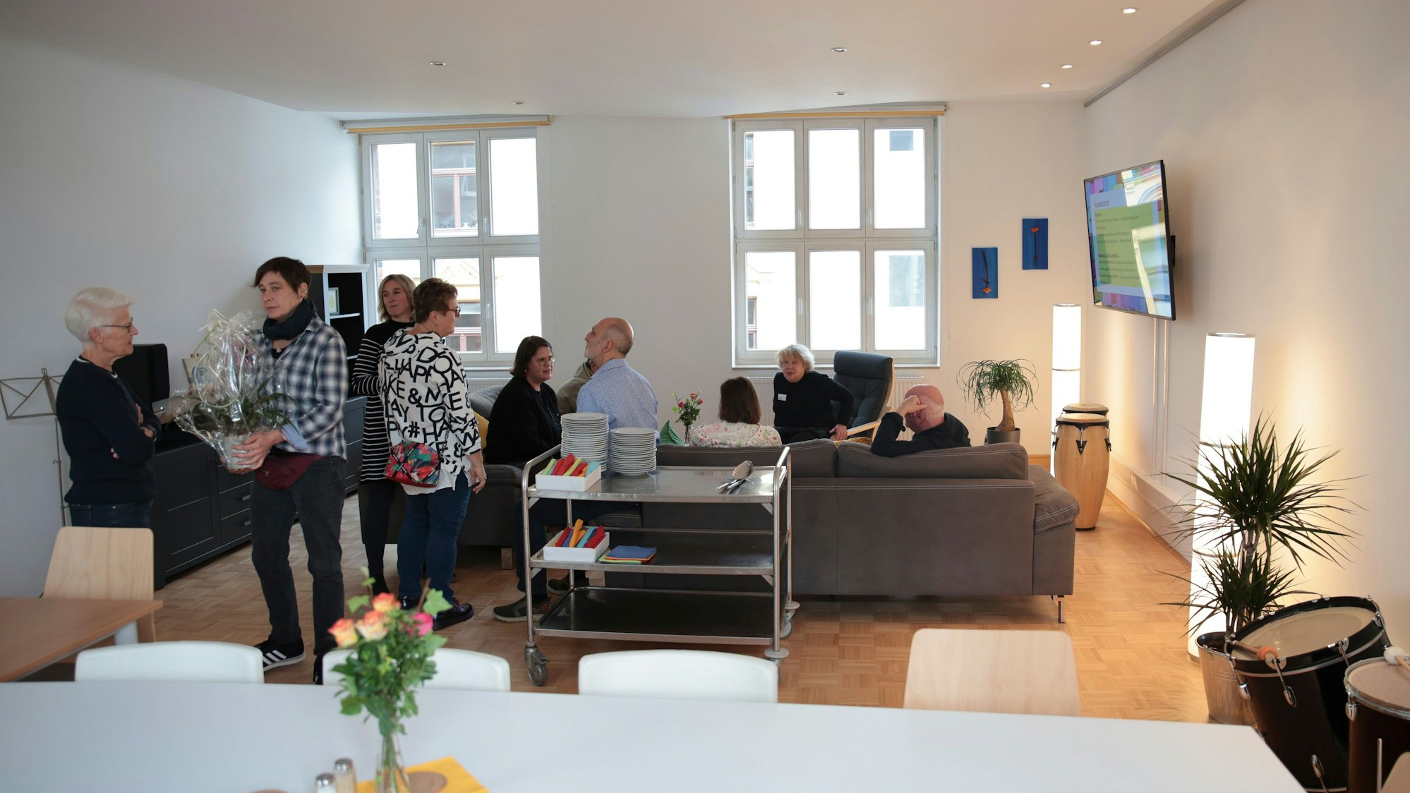 Eine Gruppe von Menschen sitzt auf Sofas oder steht daneben. An der Wand hängt ein Fernseher.