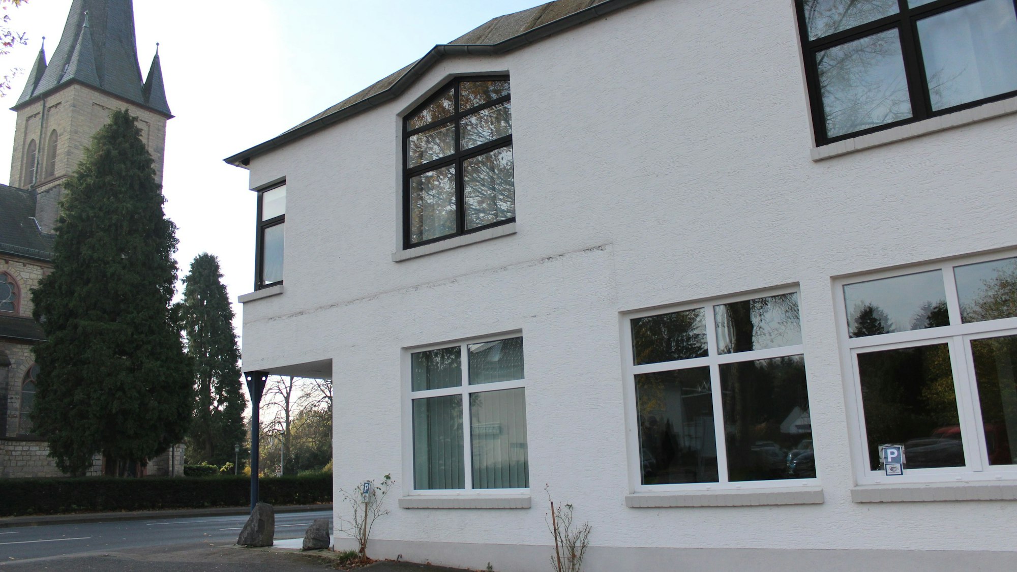 Rechts sieht man das zweistöckige Gebäude, in das die Schule einzieht. Gegenüber auf der anderen Straßenseite steht ein Kirchturm.