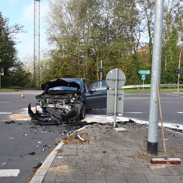 Ein an der Front vollkommen zerstörtes Auto steht auf einer Straßenkreuzung. Links im Hintergrund sind Teile eines Feuerwehrautos zu sehen. Auf der Straße liegen Splitter verteilt.