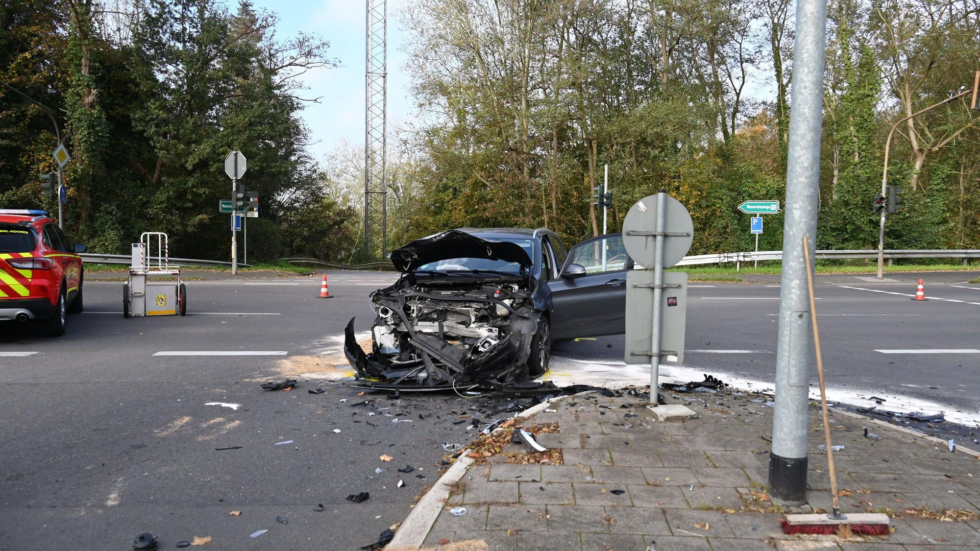 Ein an der Front vollkommen zerstörtes Auto steht auf einer Straßenkreuzung. Links im Hintergrund sind Teile eines Feuerwehrautos zu sehen. Auf der Straße liegen Splitter verteilt.