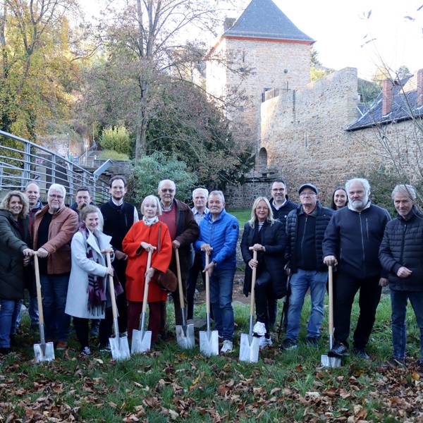 In der Nähe der Erft, im Hintergrund das Heisterbacher Tor in Bad Münstereifel, stehen Menschen mit einem Spaten in der Hand auf einer Wiese.