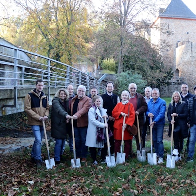 In der Nähe der Erft, im Hintergrund das Heisterbacher Tor in Bad Münstereifel, stehen Menschen mit einem Spaten in der Hand auf einer Wiese.